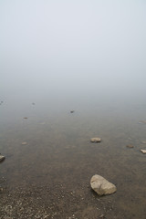 Shallow water of a lake that is lost in the fog with stones in the foreground