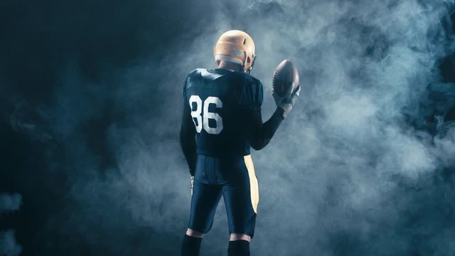 Full portrait rear view of Caucasian male American football standing with a ball, smoke background. 4K UHD RAW edited footage