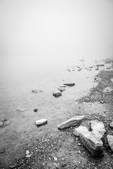 Shallow water of a lake that is lost in the fog with stones in the foreground in a black and white, high contrast tonality