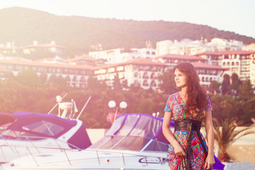 attractive brunette in long colorful dress standing alone on the beach against the backdrop of yachts and town at sunset