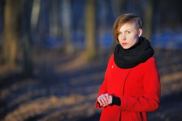 Portrait of young cute girl with a stylish haircut in a red coat and black scarf at the neck, posing in bright sun on a blurred background of trees in Park