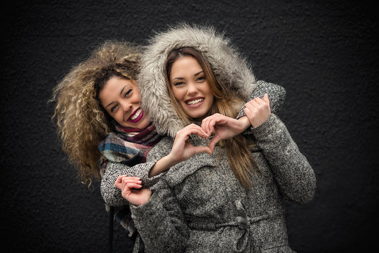Portrait of two cheerful young girlfriends embracing and creating hart shape with hands. They are looking at camera and smiling.