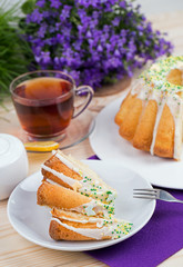 Homemade cake with icing sugar sprinkles on a plate and cup of tea on a wooden table with purple flowers in the background
