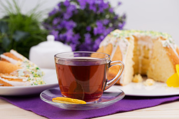 Homemade cake with icing sugar sprinkles on a plate and cup of tea on a wooden table with purple flowers in the background
