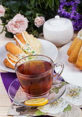 Fototapeta na wymiar Homemade cake with icing sugar sprinkles on a plate and cup of tea on a wooden table with purple and rose flowers in the background