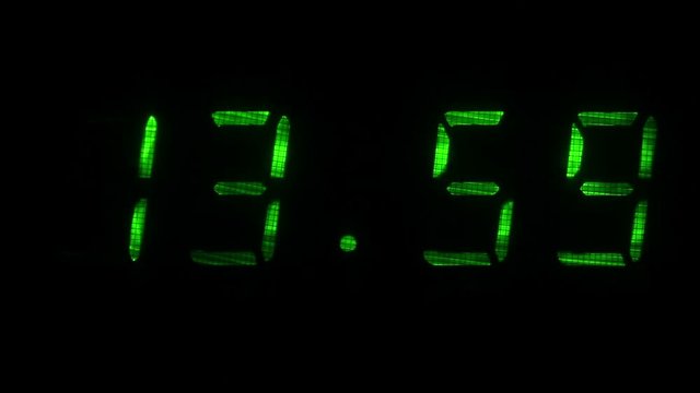 Digital clock with fluorescent display shows the time of 13 hours 59 minutes to 14 hours 00 minutes in the green on a black background