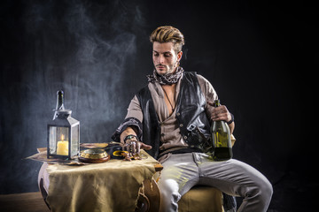 Good Looking Young Man in Pirate Fashion Outfit on Gray Background. Captured in Studio, Sitting by a Table with Bottle of Wine