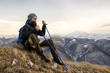 Obraz premium Young bearded hipster wearing a hat and sunglasses with Nordic walking sticks and a backpack moves in the mountains at sunset, against a background of sunset sky and volcano Elbrus