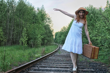 girl in a white sundress and wicker suitcase walking on rails