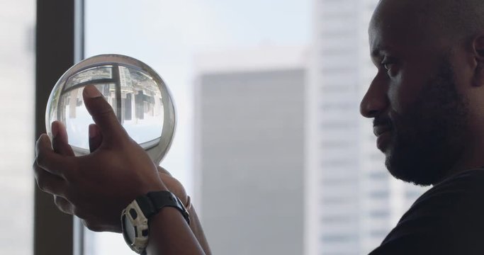Handsome African American man holds up a crystal ball and looks into it, high above Downtown Los Angeles.  Big close up, recorded hand-held in real time.