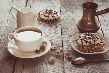White cup of coffee with milk, white milk jug, brown sugar and homemade waffle with whipped cream with decor on top. Tasty breakfast. Vintage toned.