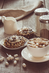 White cup of coffee with milk, marshmallow and homemade waffle with whipped cream with decor on top. Tasty breakfast. Vintage toned.