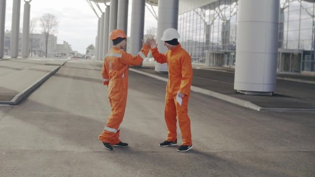 Two construction workers in orange uniform giving high five. They look very happy. New constructed building with columns at the background