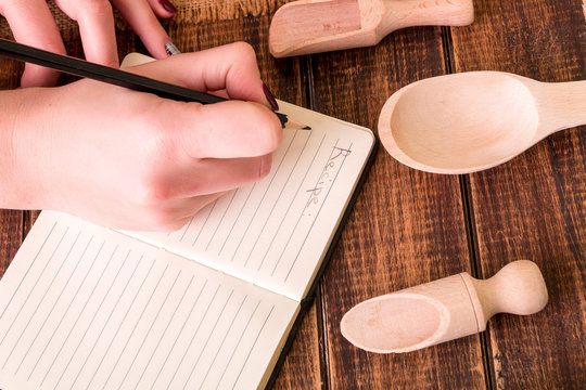 Woman hand write a recipe in cookbook. Book for recipe around utensils on wooden background.