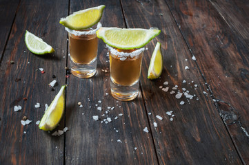 Shots of gold tequila with lime slices and salt on wood background