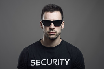 Fototapeta premium Serious moody portrait of security guard wearing sunglasses looking at camera over dark gray studio background.
