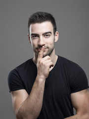 Handsome young fitness male model looking at camera with finger on lips over dark gray studio background.