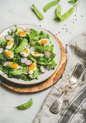 Obraz na płótnie Canvas Healthy spring green salad with radish, boiled egg, arugula, green pea and mint in white plate on olive tree wood board over grey background, selective focus