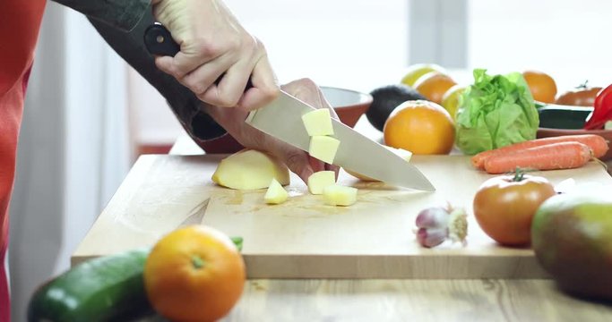 hands of man cutting yellow apple slices on brown wood plank with red and orange apron and green shirt
