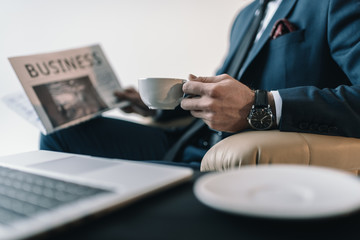 partial view of businessman with cup of coffee reading newspaper in office