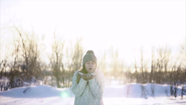 Woman wearing a warm hat with pompons of different colors, sports jacket, mittens fun with the snow on a winter day. She blows the snow that lies on her hands.