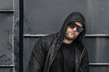 Conceptual photo of young and serious rapper in black zipped jacket and sunglasses in front of the wall. He is looking forward at something. Barcelona, Spain.