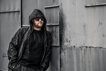 Conceptual photo of young and serious rapper in black zipped jacket and sunglasses in front of the wall. He is looking forward at something. Barcelona, Spain.