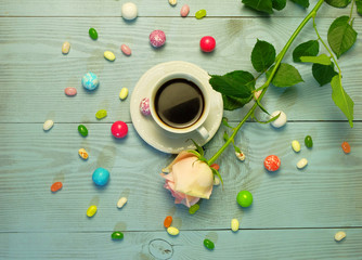 A cup of coffee, colorful candies and jelly beans and a rose on a blue wooden table.