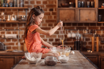 'side view of girl sitting on table and mixing ingredients for cake