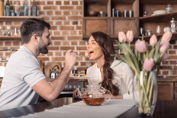 'side view of smiling man feeding woman with cake in kitchen