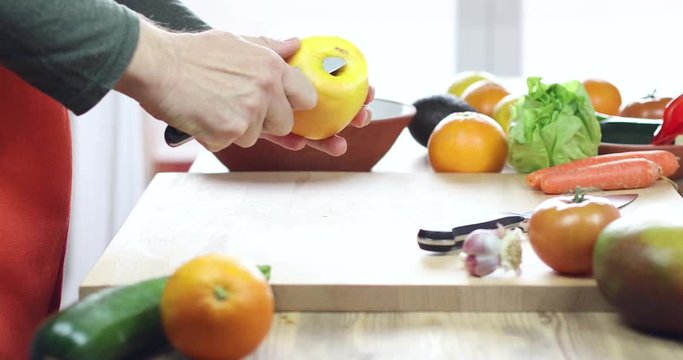 hands of man cutting garlic slices on brown wood plank with red and orange apron and green shirt
