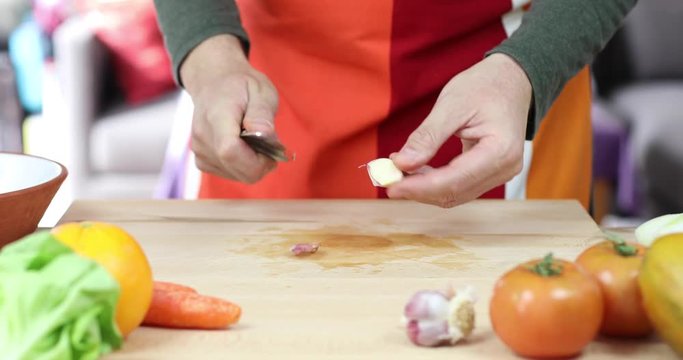 hands of man cutting garlic slices on brown wood plank with red and orange apron and green shirt
