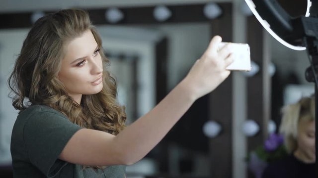 Pathos girl with painted eyebrows, eyeliner let down for centuries, curls on the hair makes selfie on her mobile phone. Girl posing to get better in pictures