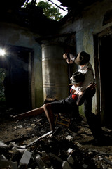 Conceptual photo of murderer and victim. Young woman is sitting on a chair with bound hands and man is standing next to her with gun in hands. They are dressed in retro style. Ruined place.