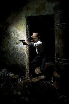 Portrait of a gangster dressed in retro suit with hat, smoking cigar and holding gun in hands. Ruined place.
