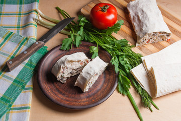Two pieces of pita bread or lavash roll with cottage cheese or curd, chicken, tomatoes and herbs - dill, onion, parsley on brown clay plate..