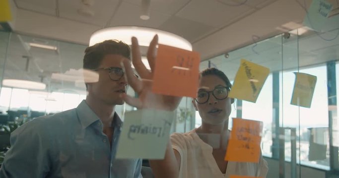 Business colleagues brainstorming together at a glass wall wth adhesive notes. Businesswoman with male coworker discussing new ideas.