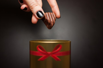 mano che tiene caramella di cioccolato della donna, tirato fuori da una scatola d'oro rotonda con un fiocco rosso su sfondo nero