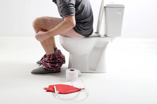 Mężczyzna siedzi na sedesie z leżącym obok wlewnikiem do lewatywy 