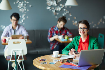 Young creative businesswoman wearing glasses and colorful casual clothes smiling looking at camera while working with laptop at table in cafe, two other people in background