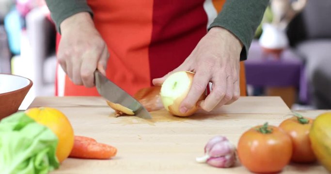 hands of man cutting onion slices on brown wood plank with red and orange apron and green shirt
