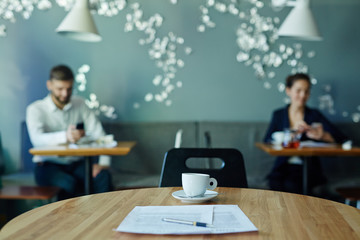 Image of business documents on empty table in cafe with two businesspeople working in background during coffee break