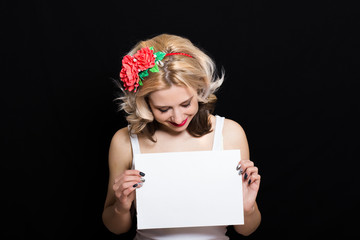 Woman with blonde curls and red lipstick at the rim with red flowers holding a white sheet on a black background and smiling. Close-up. Space for text