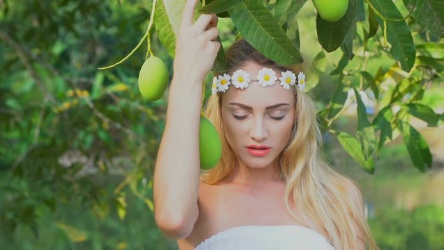 Closeup face of stunning blond woman in white dress and wreath of white flowers looking at the camera and posing with mango fruit tree in the garden over lake background - video in slow motion