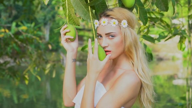 Closeup face of stunning blond woman in white dress and wreath of white flowers looking at the camera and posing with mango fruit tree in the garden over lake background - video in slow motion