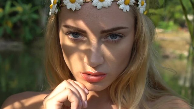 Closeup face of stunning blond woman in wreath of white flowers and light makeup looking at the camera and posing near mango fruit tree in the garden over lake background