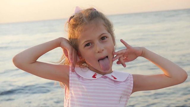 beautiful small girl grimaces and shows tongue and teeth on the background of the sunset sea