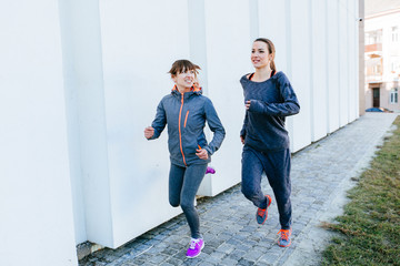 Obraz premium Two sports women in gray sportswear running beside white wall in sunny spring morning