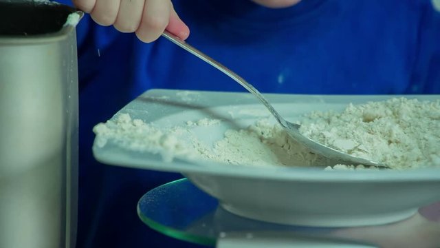 A toddler is putting many spoons of flour into a different container. He is playing by himself in the kitchen.
