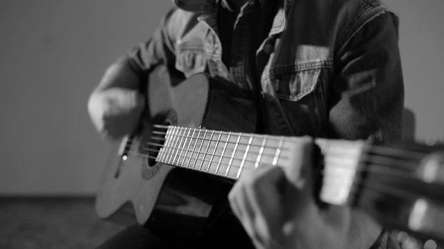 Male guitarist playing on acoustic guitar sitting at gray wall background - monochrome